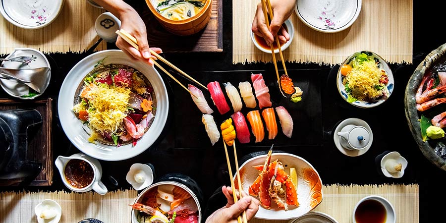 A Japanese meal fills a table and three people reach over the food to grab some with their chopsticks.